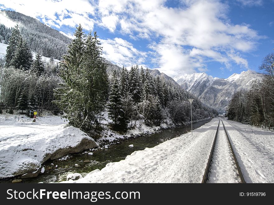 Snow covered railroad tracks next to stream in Alps through pine forest on sunny day. Snow covered railroad tracks next to stream in Alps through pine forest on sunny day.