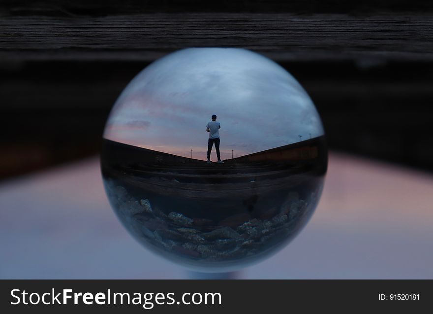 Glass sphere illuminated from above, and with the small reflected image of a man wearing shorts in it, upper background dark and lower part light gray. Glass sphere illuminated from above, and with the small reflected image of a man wearing shorts in it, upper background dark and lower part light gray.