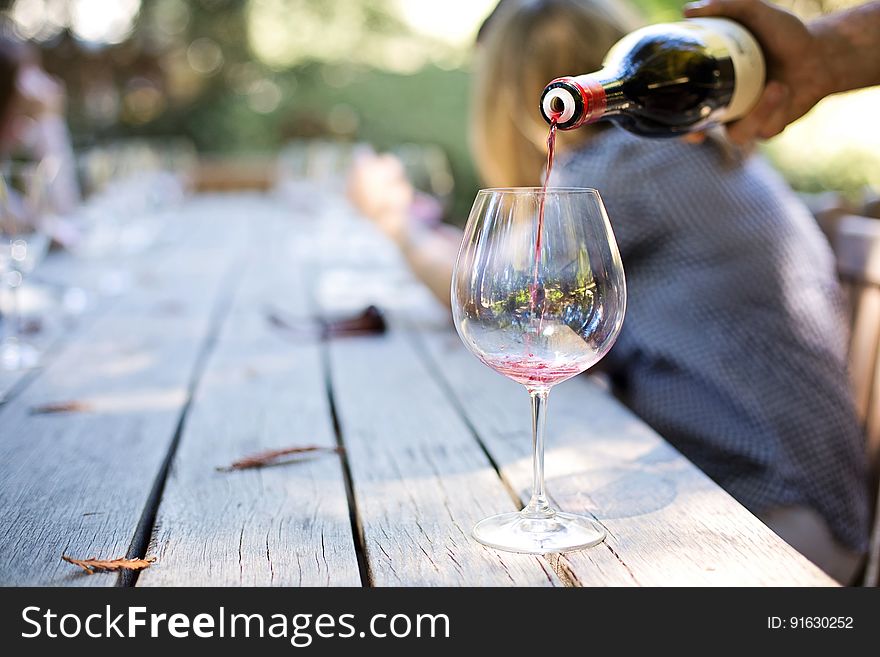 A person pouring red wine in a glass on desk with people in the background. A person pouring red wine in a glass on desk with people in the background.