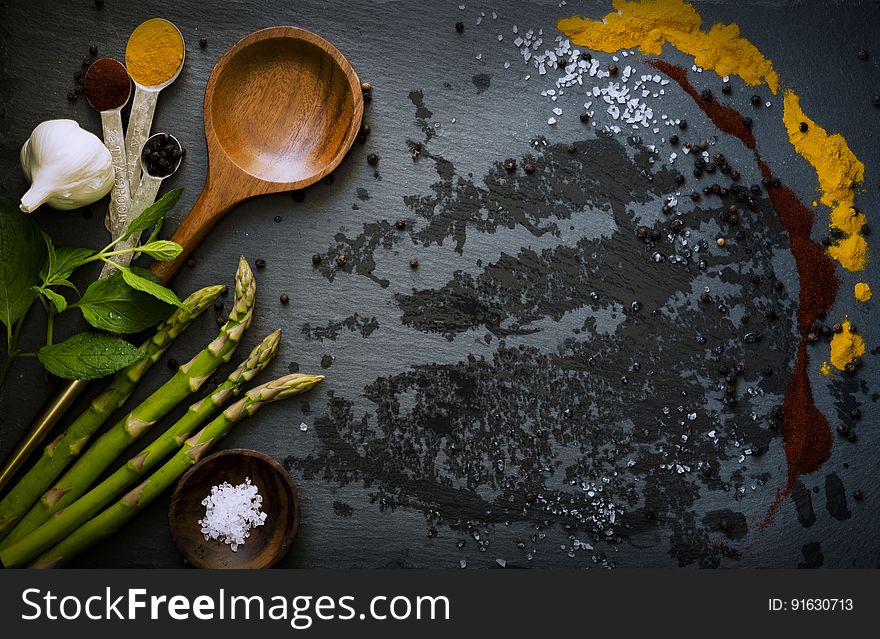 Food ingredients laid out on a dark surface including asparagus, garlic clove, mint leaves, herbs and paprika alongside spoons and a small bowl. Food ingredients laid out on a dark surface including asparagus, garlic clove, mint leaves, herbs and paprika alongside spoons and a small bowl.