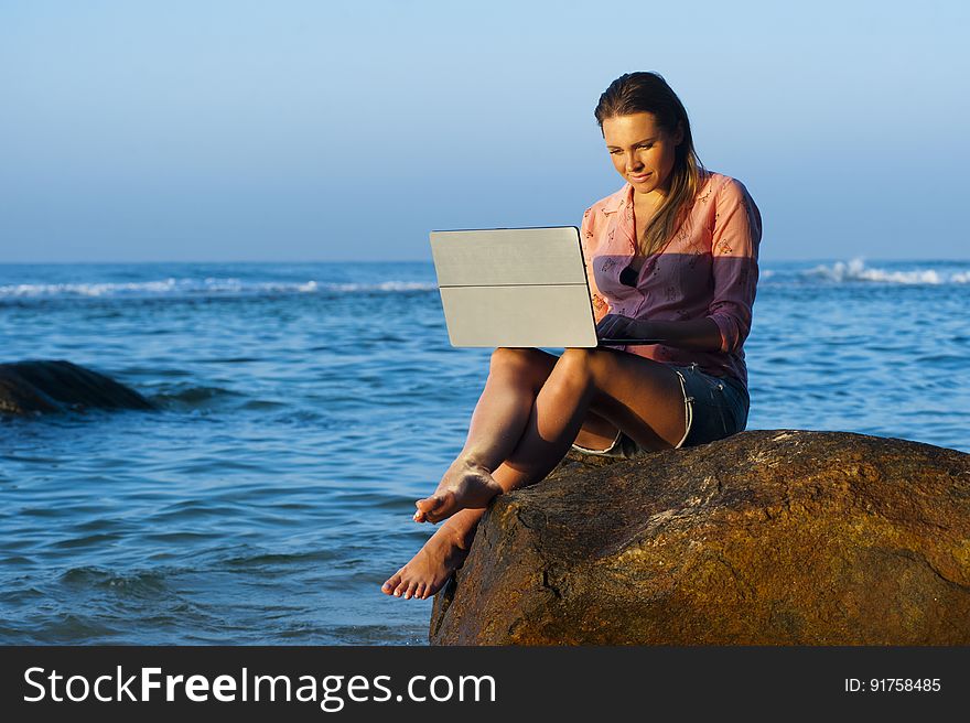 A woman working on a laptop while sitting on a rock at the seaside. A woman working on a laptop while sitting on a rock at the seaside.