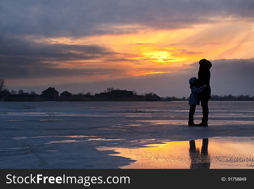 Silhouetted mother and child by frozen lake at sunset.