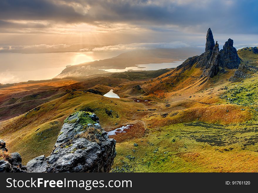 Rolling landscape with Autumn colors, shades of green and orange, with needle like rocks, pale blue lake, and blue gray sky. Rolling landscape with Autumn colors, shades of green and orange, with needle like rocks, pale blue lake, and blue gray sky.