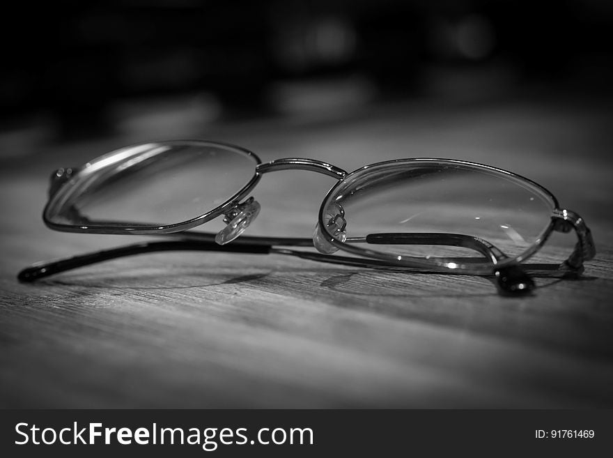 A black and white image of eyeglasses on a wooden desk. A black and white image of eyeglasses on a wooden desk.