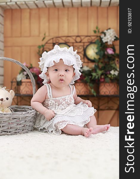 Baby Wearing White Headband and White Lace Floral Dress Sitting Beside Gray Wicker Basket
