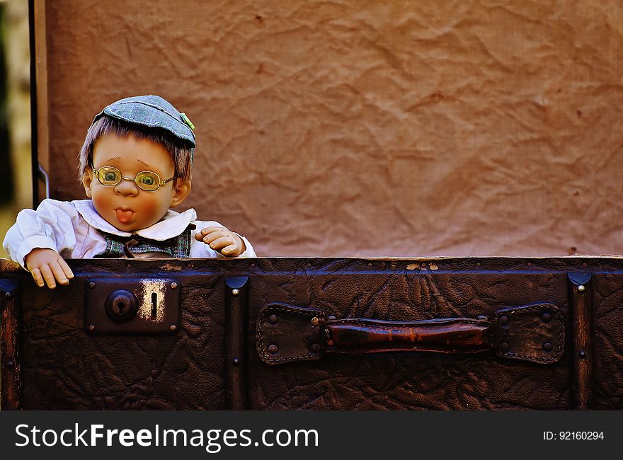 Antique baby boy (doll) with rosy cheeks dressed in white jacket and blue flat cap wearing spectacles standing behind a leather suitcase, mostly brown textured background. Antique baby boy (doll) with rosy cheeks dressed in white jacket and blue flat cap wearing spectacles standing behind a leather suitcase, mostly brown textured background.