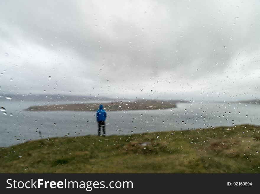 Distant shot of the back of a man standing on the shore by water on a cloudy, foggy day. Focus of photo is mist on lens of the camera with shallow depth of field. Distant shot of the back of a man standing on the shore by water on a cloudy, foggy day. Focus of photo is mist on lens of the camera with shallow depth of field.