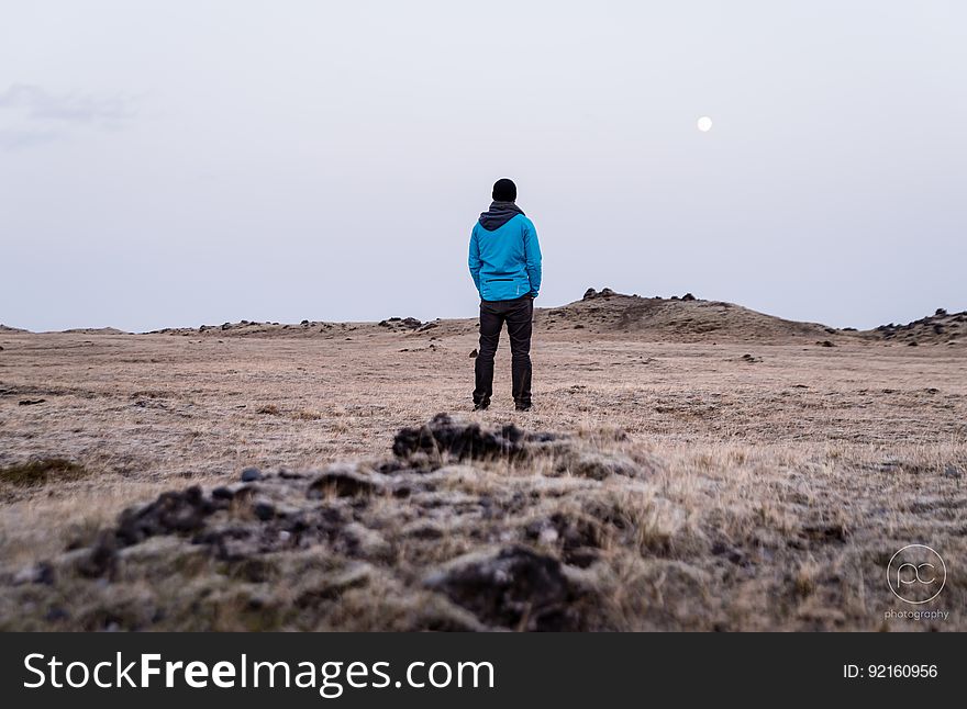 Rear portrait of man in blue jacket standing in bare field with moon in sky. Rear portrait of man in blue jacket standing in bare field with moon in sky.