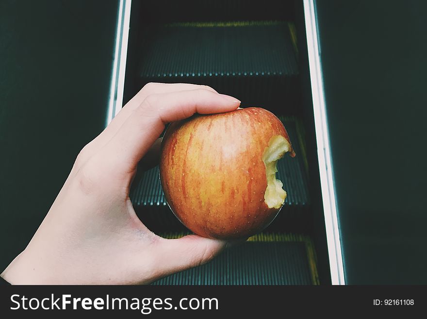 A person holding an apple with a bite taken out of it on escalators. A person holding an apple with a bite taken out of it on escalators.