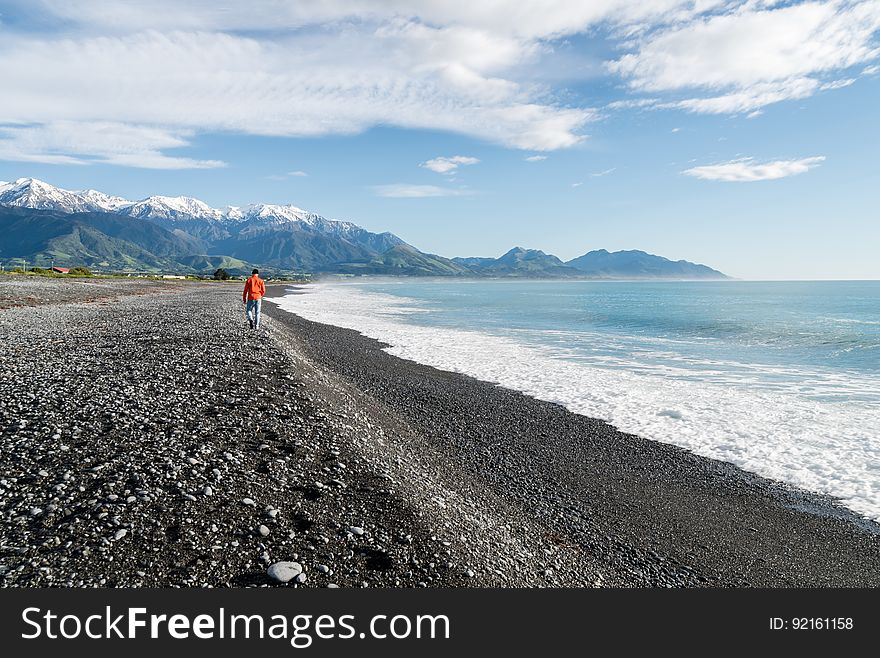 A pebble beach with a person walking next to the water with mountains in the distance.