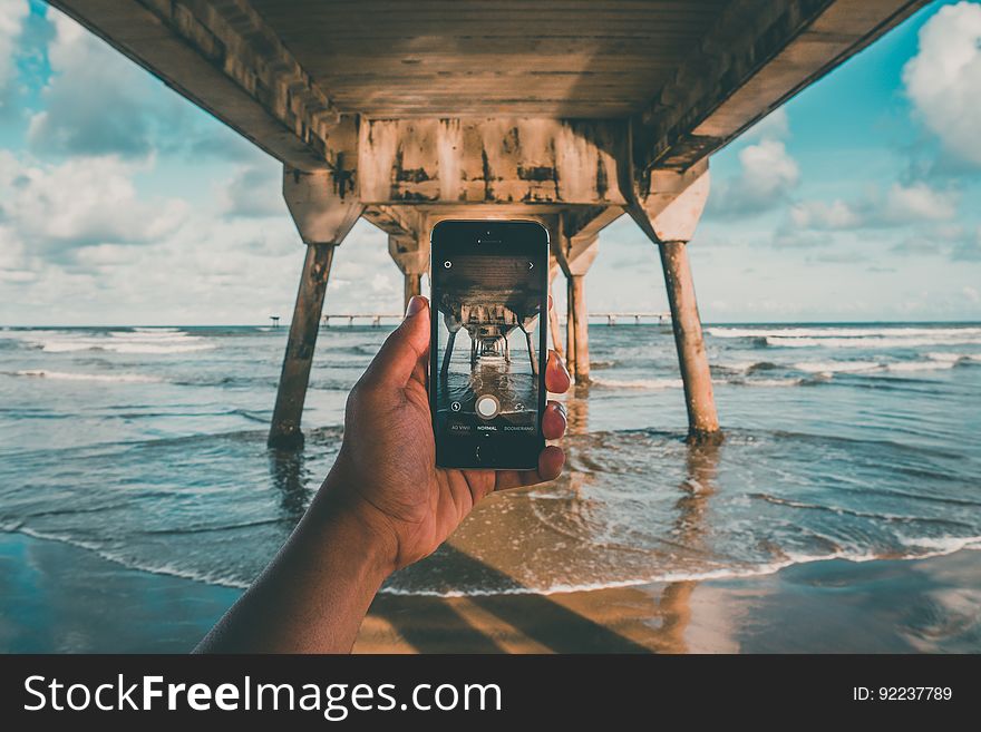 Hand holding mobile phone to take a picture while the person is in the sea under a wooden jetty on legs, background pale blue sky and clouds. Hand holding mobile phone to take a picture while the person is in the sea under a wooden jetty on legs, background pale blue sky and clouds.