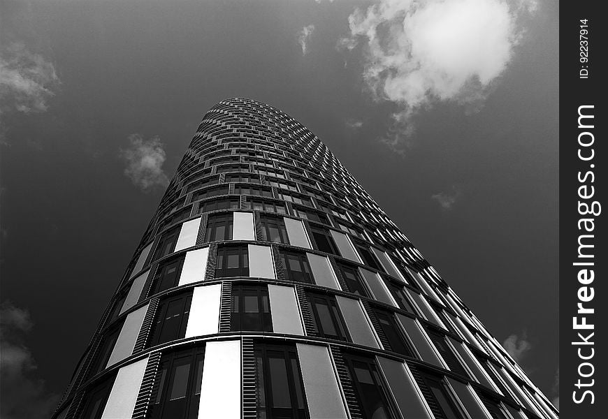 Unusual building, skyscraper built of concrete, steel and glass but with curved facade and seen from (above) ground pointing upwards, gray sky and cloud. Unusual building, skyscraper built of concrete, steel and glass but with curved facade and seen from (above) ground pointing upwards, gray sky and cloud.