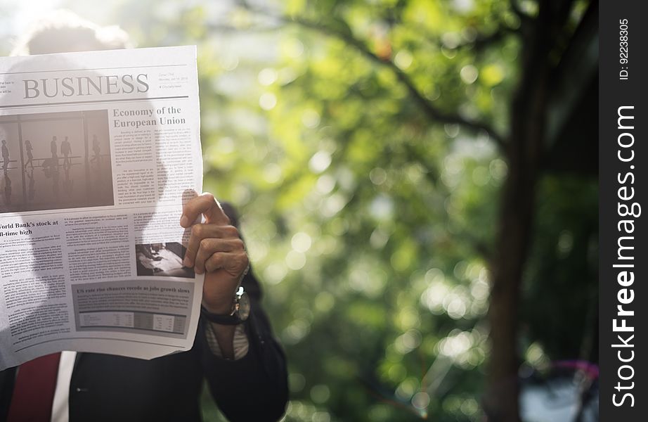 Man in a business suit reading the business section of a newspaper outdoors by trees, with his face obscured and his silhouette visible on the newspaper from the sunlight behind. Man in a business suit reading the business section of a newspaper outdoors by trees, with his face obscured and his silhouette visible on the newspaper from the sunlight behind.