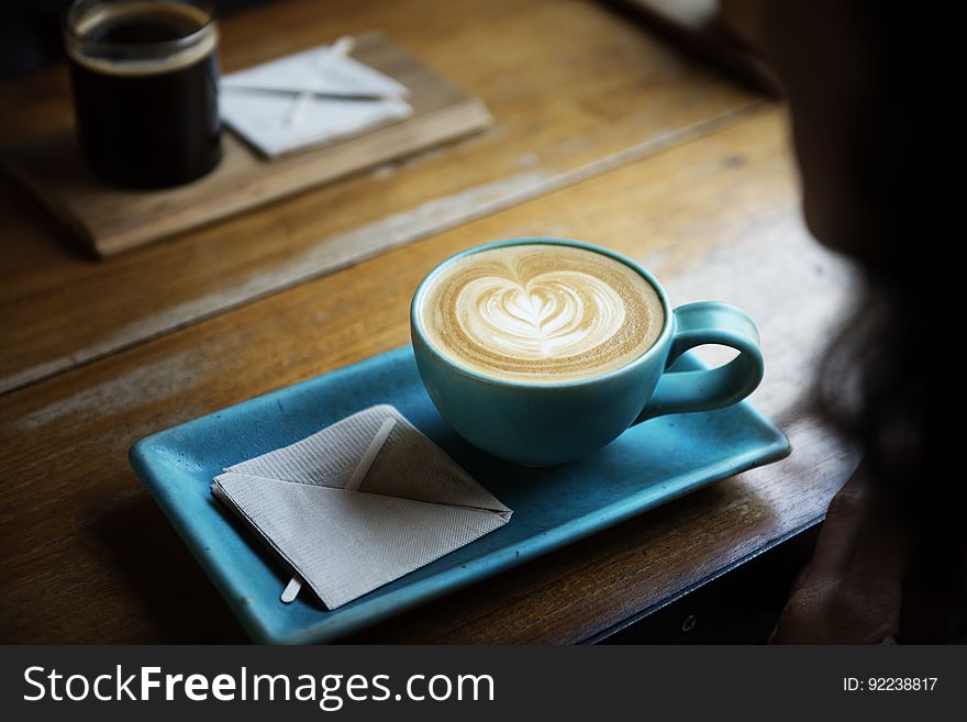 wooden table with cup of coffee in stylish matching blue rectangular shaped saucer with heart shape in foam on the top and napkin square in the saucer. wooden table with cup of coffee in stylish matching blue rectangular shaped saucer with heart shape in foam on the top and napkin square in the saucer.