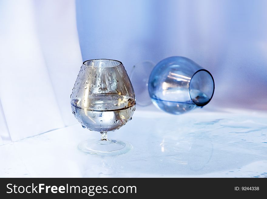 Two wineglasses, one stands the second lies,  on wet glass. Both with a liquid.
