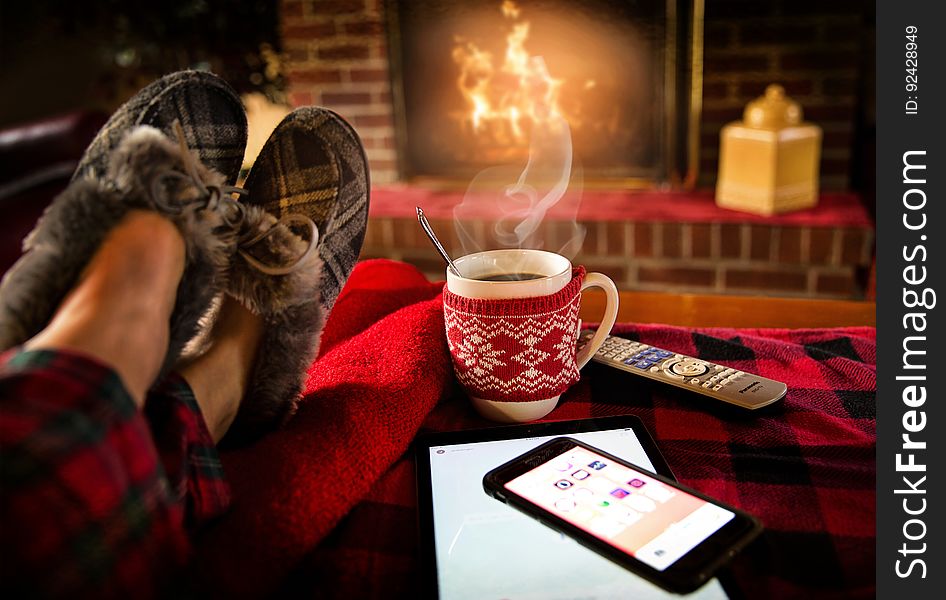 A person with slippers relaxing by the fireplace with a cup of coffee and smartphone. A person with slippers relaxing by the fireplace with a cup of coffee and smartphone.