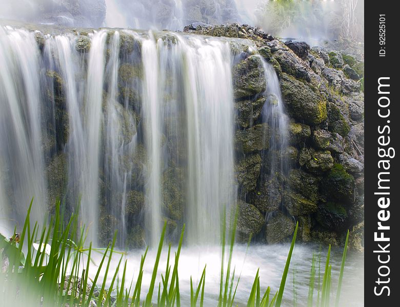 Time lapse scenic view of waterfall over rocky cliff with blades of grass in foreground.