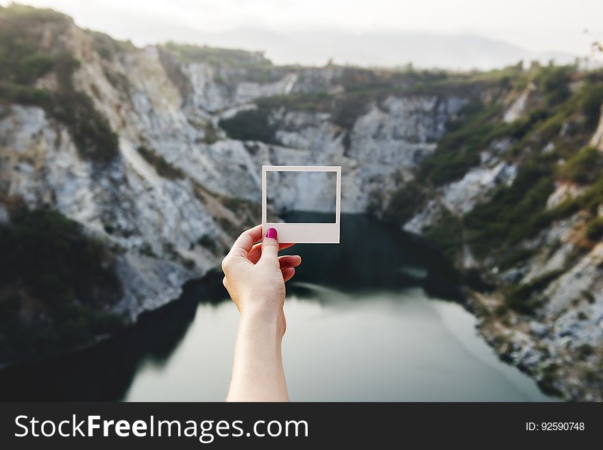 A person holding a polaroid photo that captures the cliffs in front of it. A person holding a polaroid photo that captures the cliffs in front of it.