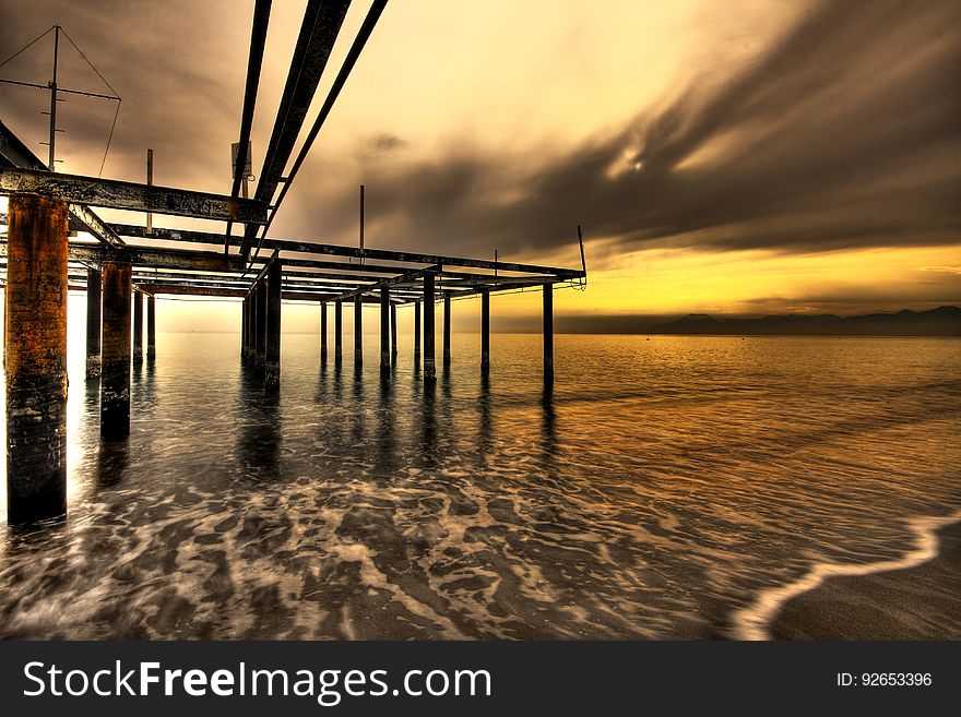 Pier structure without the usual wooden boards lit by an orange and yellow light of the sunset with the tide coming in and the sea decorated by a foam pattern. Pier structure without the usual wooden boards lit by an orange and yellow light of the sunset with the tide coming in and the sea decorated by a foam pattern.