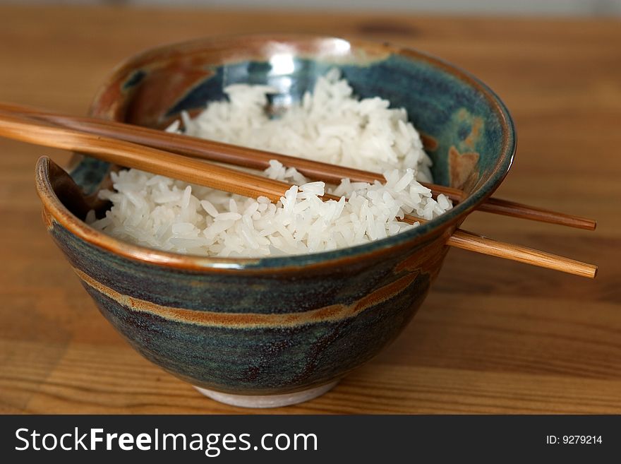 A bowl of steamed rice with wooden chopsticks through holes of the bowl on a wooden surface. A bowl of steamed rice with wooden chopsticks through holes of the bowl on a wooden surface.