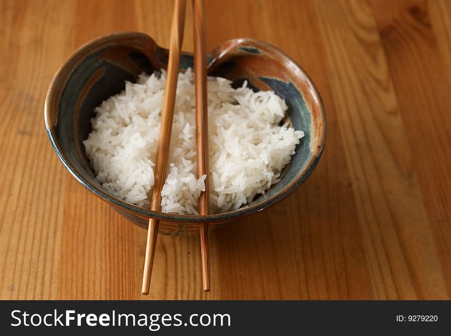 A bowl of steamed rice with wooden chopsticks through holes of the bowl on a wooden surface. A bowl of steamed rice with wooden chopsticks through holes of the bowl on a wooden surface.