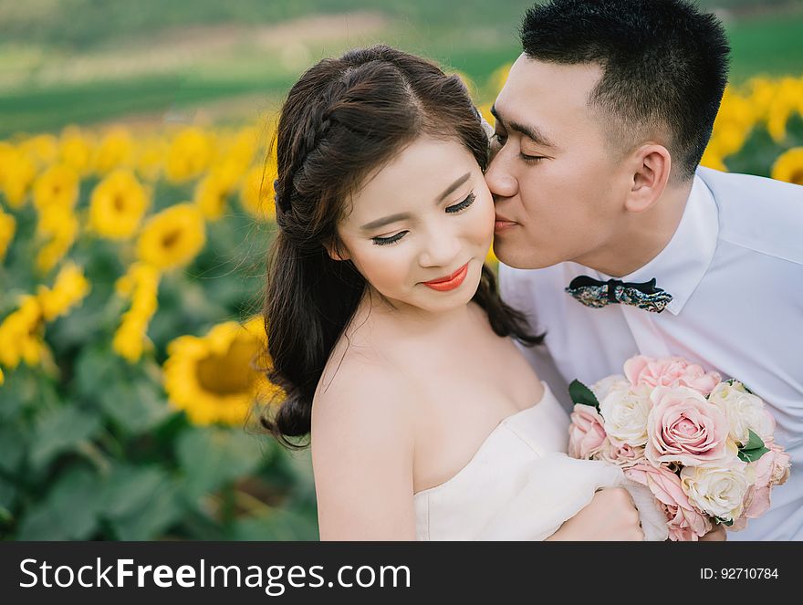 Wedding picture of beautiful bride in white wedding dress holding posy of pink roses being kissed by an adoring groom with a unique background of blurred yellow sunflowers. Wedding picture of beautiful bride in white wedding dress holding posy of pink roses being kissed by an adoring groom with a unique background of blurred yellow sunflowers.