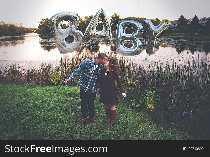 Loving couple standing beside a river kissing passionately with rubber balloons over their heads spelling "baby" in uppercase letters ( 3D shapes) possibly indicating the desire for a child. Loving couple standing beside a river kissing passionately with rubber balloons over their heads spelling "baby" in uppercase letters ( 3D shapes) possibly indicating the desire for a child.
