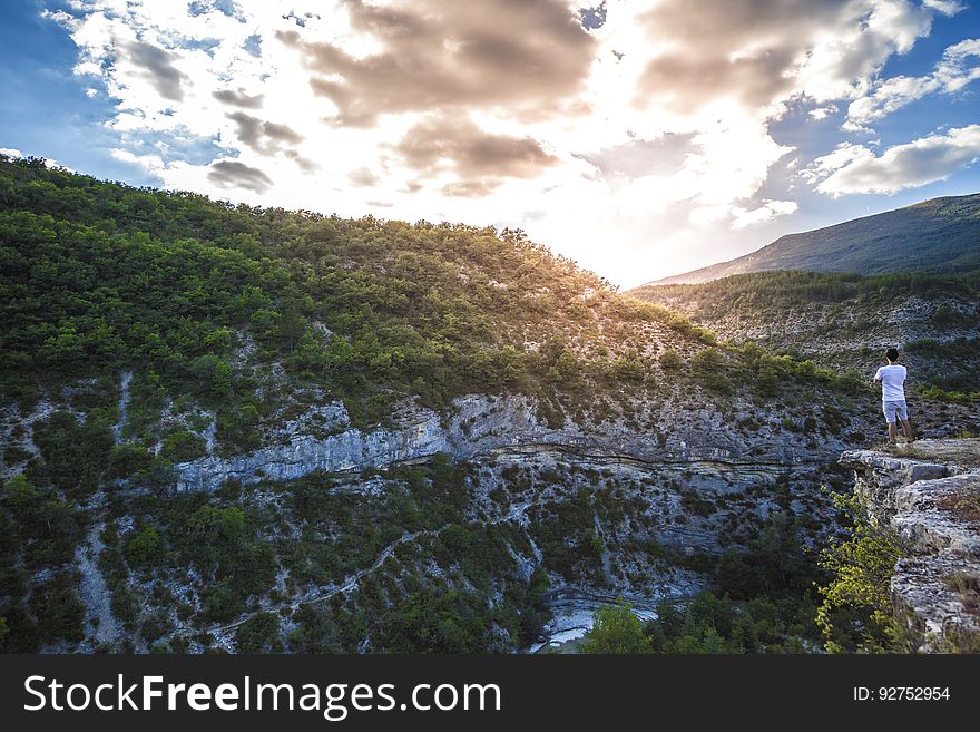 Man in White Shirt Standing on Top of Mountain Cliff Under Cumulus Clouds