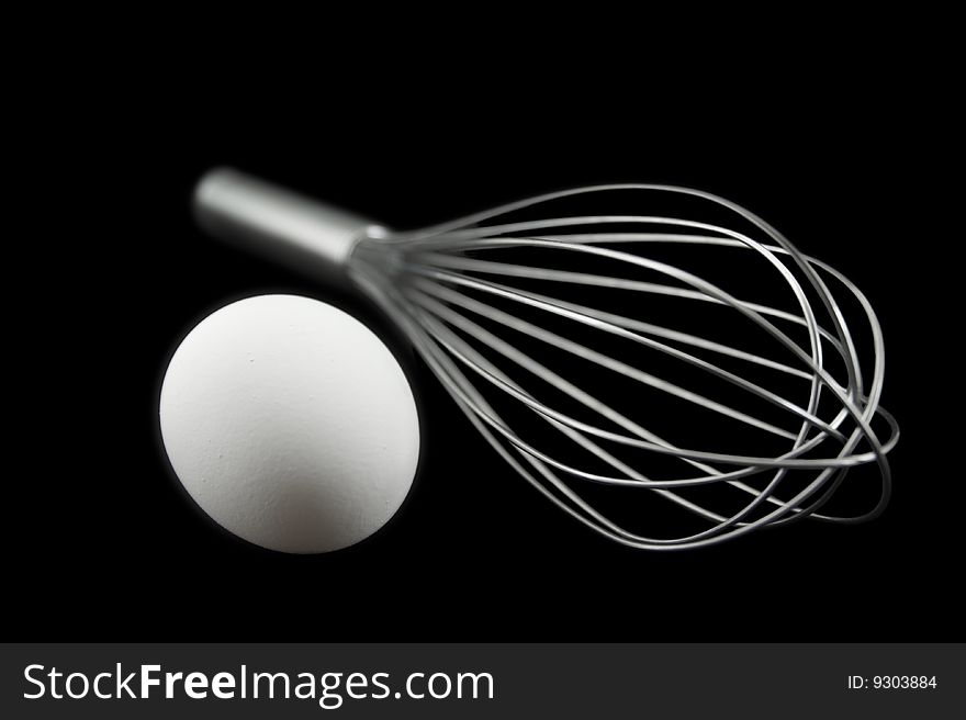 A white egg next to a soft focus steel wisk isolated on a black black background. A white egg next to a soft focus steel wisk isolated on a black black background