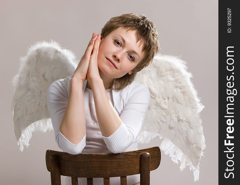 Silhouette of a pensive girl in white clothes on the light background sitting on the chair with wings of an angel behind her back. Silhouette of a pensive girl in white clothes on the light background sitting on the chair with wings of an angel behind her back