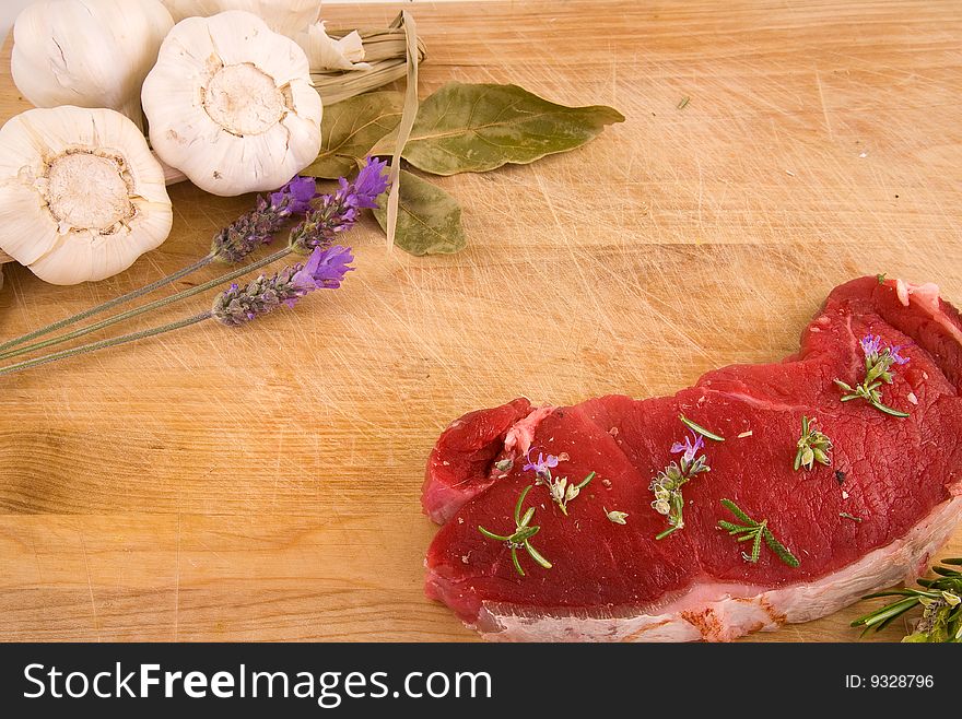 Two beef steaks, and a set of Mediterranean ingredients over a wooden board ready to be chopped and used to flavour a meal. Two beef steaks, and a set of Mediterranean ingredients over a wooden board ready to be chopped and used to flavour a meal