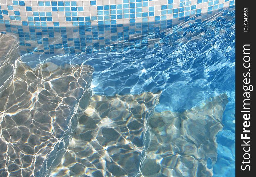 An image of steps going into a swimming pool with mosaic tiles around the edge. An image of steps going into a swimming pool with mosaic tiles around the edge