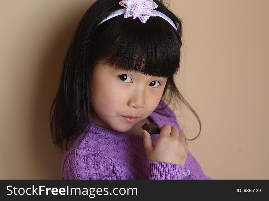 Asian Chinese Girl eating a small piece of chocolate with a head band with bow on top and purple sweater. Asian Chinese Girl eating a small piece of chocolate with a head band with bow on top and purple sweater