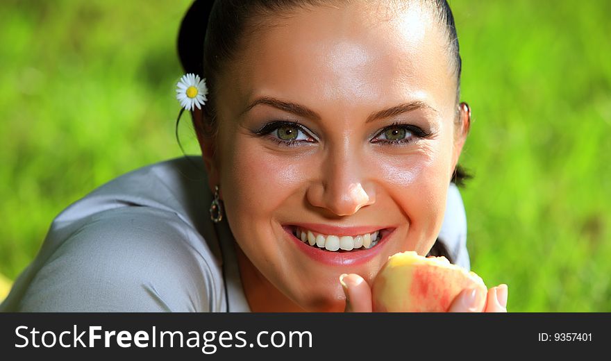 Attractive young girl is eating a fresh peach. She is lying on a nice fresh green grass meadow and smiling with a flower behind her ear. Attractive young girl is eating a fresh peach. She is lying on a nice fresh green grass meadow and smiling with a flower behind her ear