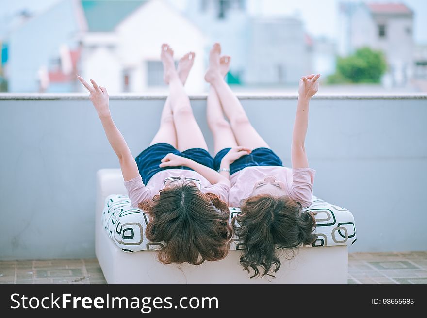 Two female friends in similar clothes laying side by side outside on a deck with their legs propped and crossed on a low wall away from the viewer, each girl raising two fingers in a peace or victory sign. Two female friends in similar clothes laying side by side outside on a deck with their legs propped and crossed on a low wall away from the viewer, each girl raising two fingers in a peace or victory sign.