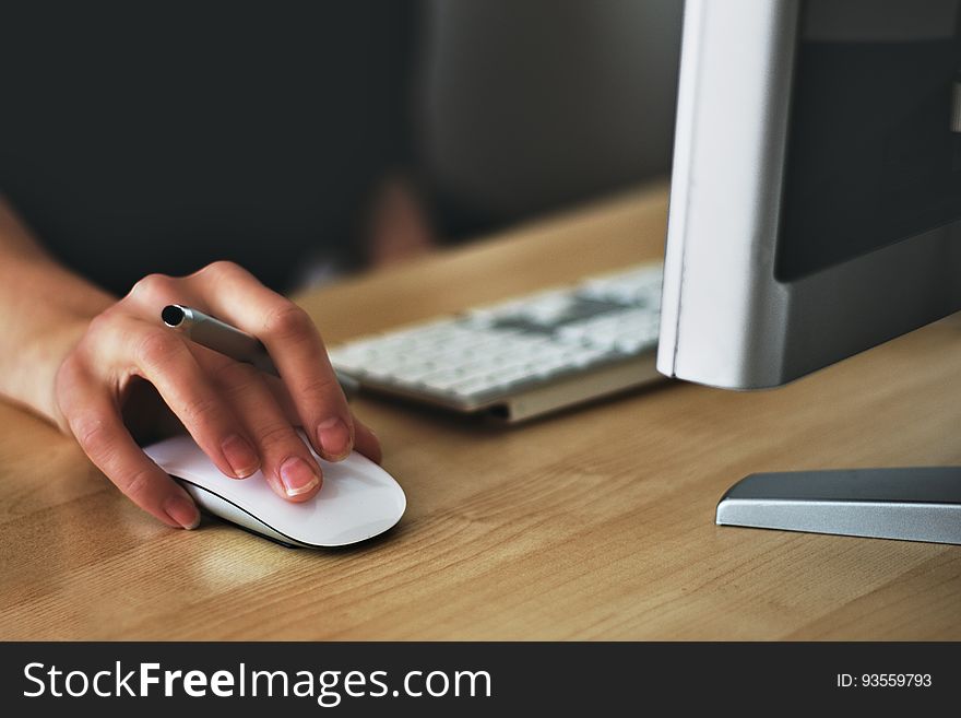 Selective focus on woman's hand and fingers on mouse next to computer keyboard and display, also holding pen between forefinger and second finger. Selective focus on woman's hand and fingers on mouse next to computer keyboard and display, also holding pen between forefinger and second finger.