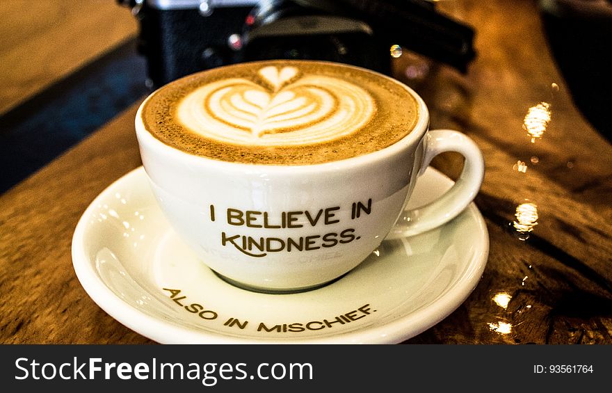 White chine cup and saucer with message I Believe in Kindness and Also in Mischief with latte coffee drink. White chine cup and saucer with message I Believe in Kindness and Also in Mischief with latte coffee drink.