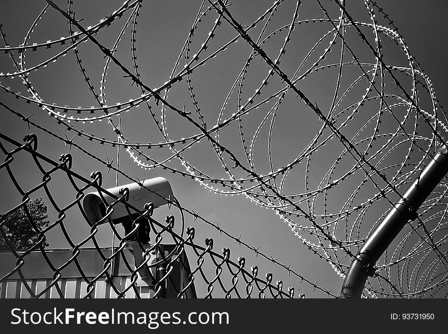 A fence with razor wire on top and a CCTV camera behind it. A fence with razor wire on top and a CCTV camera behind it.