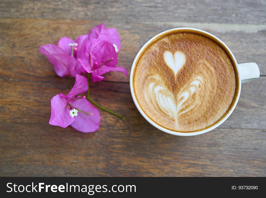 Overhead of cup of latte with heart shapes in froth with purple flowers on wooden tabletop. Overhead of cup of latte with heart shapes in froth with purple flowers on wooden tabletop.