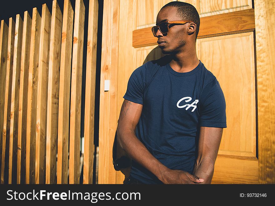 Portrait of African man in blue t-shirt wearing sunglasses leaning against wooden wall in sunshine. Portrait of African man in blue t-shirt wearing sunglasses leaning against wooden wall in sunshine.