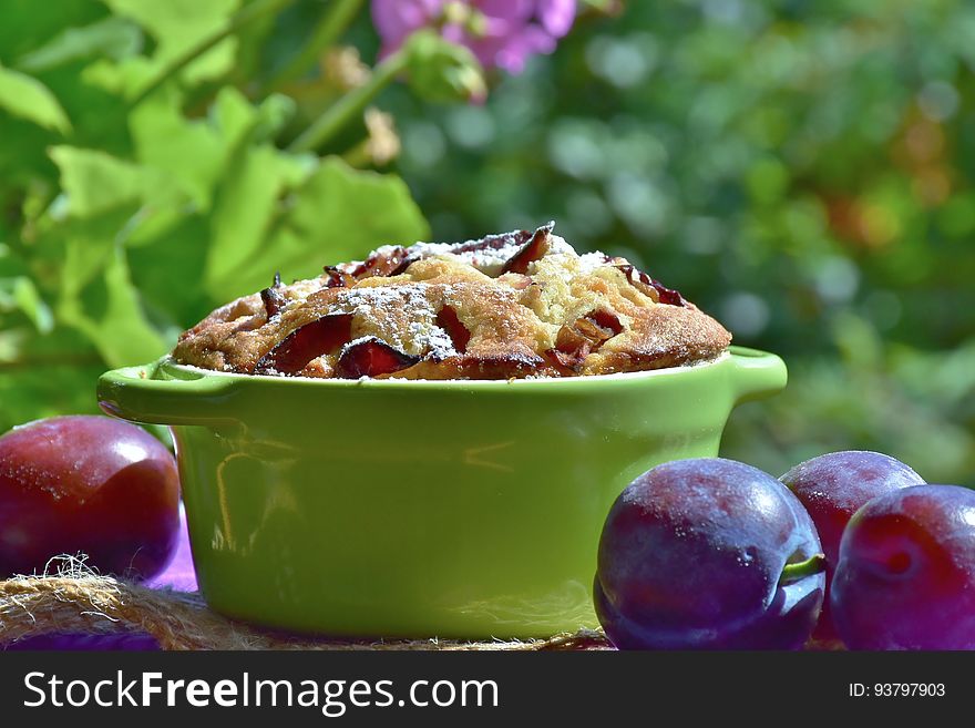 Baked cake with fresh plumbs on table outdoors, green leaves and flowers in background.