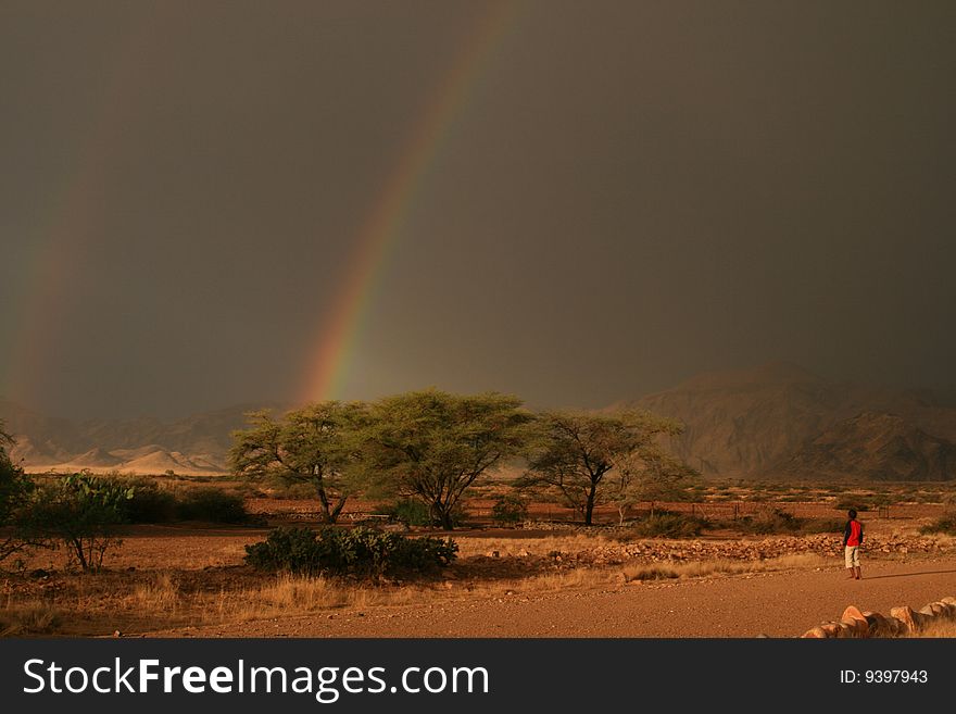 African boy watching a dark sky with beautiful rainbow full with colors in the namib naukluft park, Namibia. African boy watching a dark sky with beautiful rainbow full with colors in the namib naukluft park, Namibia.