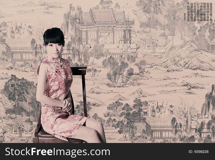 A Chinese girl in traditional dress sits in front of a Chinese old painting. Chinese characters on the picture are China's ancient poesy. Usually there is a piece of poesy in the old painting above of China. Adjust the tone on later stage in picture.