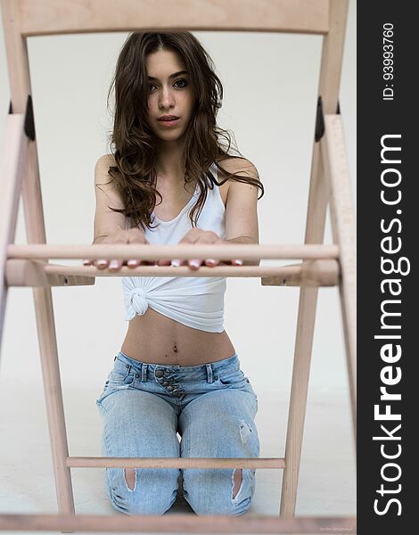Brunette girl in white top and jeans sitting on his knees in front of the wooden chair. Vertical photo