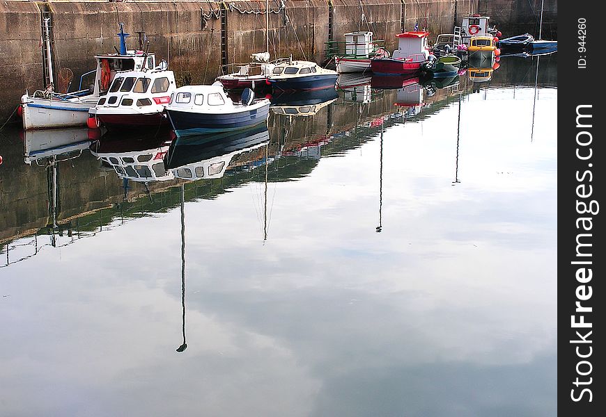 One side of the harbour with the summer season boats and yawls reflected in the calm still water with the sky. One side of the harbour with the summer season boats and yawls reflected in the calm still water with the sky.