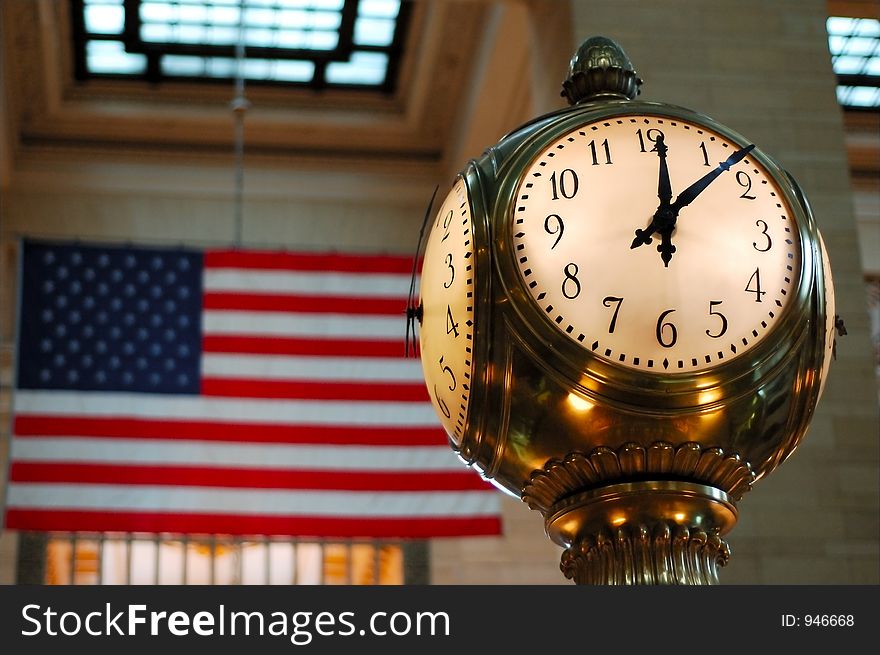 Clock on Information Booth at Grand Central Station New York City. The American flag is blurred in the background. The time is just past high noon. Clock on Information Booth at Grand Central Station New York City. The American flag is blurred in the background. The time is just past high noon