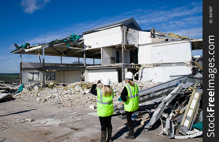 Two women in flourescent jackets watch a building being demolished. Two women in flourescent jackets watch a building being demolished