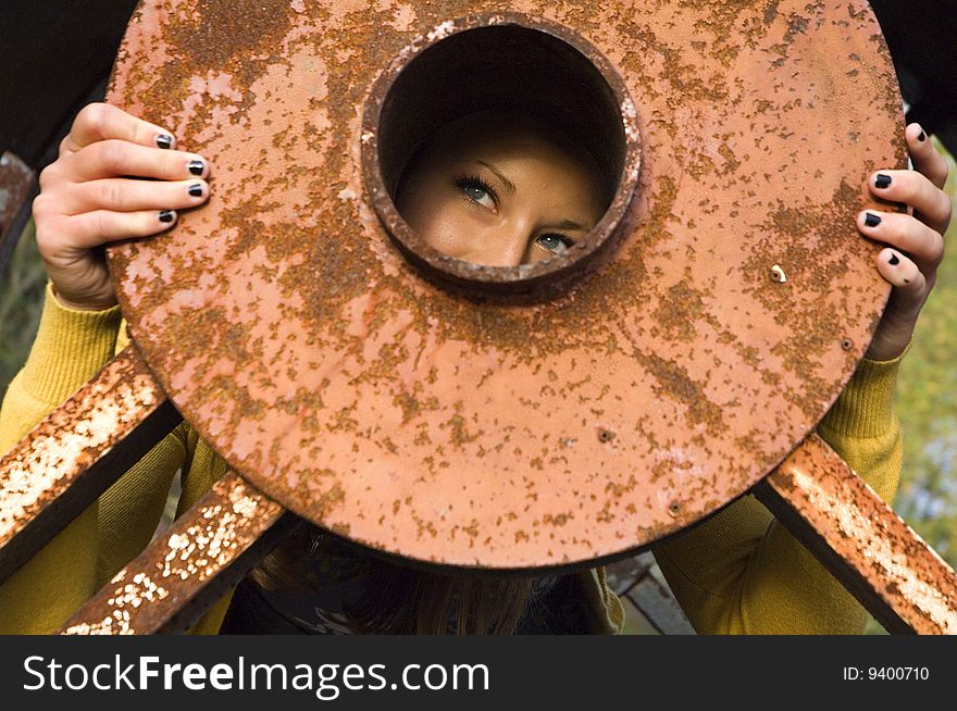 Beautiful young woman peers through a large orange rusty metal disc. Beautiful young woman peers through a large orange rusty metal disc.
