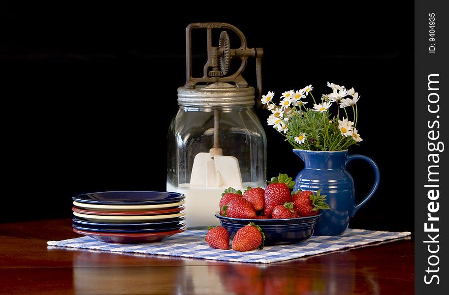 Fresh strawberries in cobalt blue bowl, colorful plates stacked to the side and antique butter churn filled with cream in background. Fresh strawberries in cobalt blue bowl, colorful plates stacked to the side and antique butter churn filled with cream in background