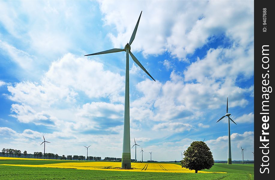Wind turbines among rapeseed field and green meadows against a cloudy blue sky. Wind turbines among rapeseed field and green meadows against a cloudy blue sky